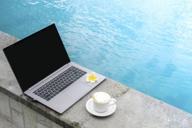Yüzme havuzunda bir fincan kahveyle dizüstü bilgisayar, tatilde serbest çalışan bir işçide çalışma alanı.