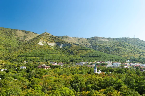 Housing estate at the foot of the Caucasus Mountains. Green hills covered with trees, residential buildings, worship cross on top of hill.