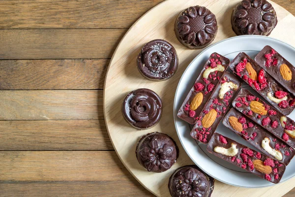 Healthy homemade recipe chocolate candies and sticks with almonds, cashews and raspberries, lie on white plate on round kitchen board. Copy space. Top view. Round tasty sweets on a wooden background