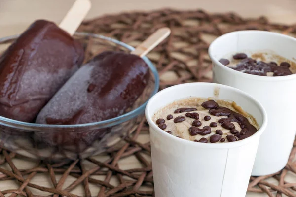 Raw banana ice cream with coconut cream, coated with natural chocolate, in a white paper cup, and chocolate popsicles on sticks in glass bowl. Healthy vegan dessert. Harmless homemade sweets
