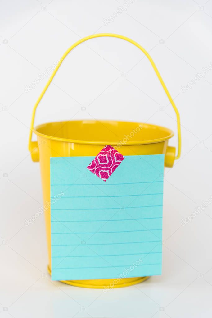 Aqua colored lined paper list taped to front of bright yellow miniature bucket with solid white background to illustrate bucket list