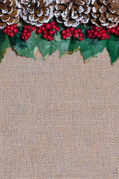 Border of Christmas pinecones, holly leaves and red berries on rustic burlap fabric background