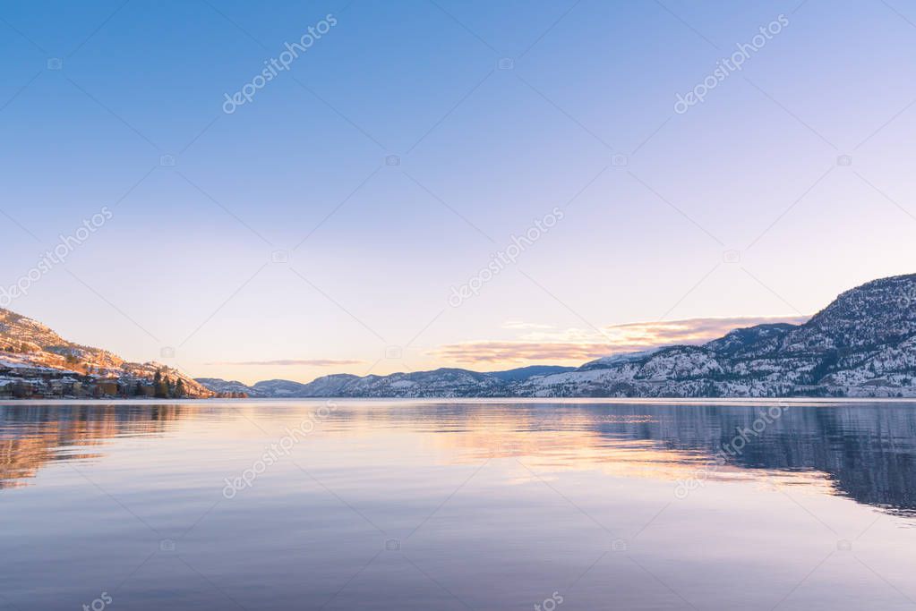 Clear sky with sunset colors reflected in calm lake waters, with view of distant snow covered mountains