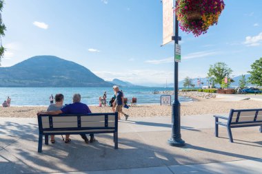 Penticton, British Columbia/Kanada - 22 Haziran 2019: İnsanlar banklarda oturup Lakeshore Drive boyunca yürür ler ve yaz turizm sezonunda Okanagan Gölü'ndeki plajın keyfini çıkarırlar..