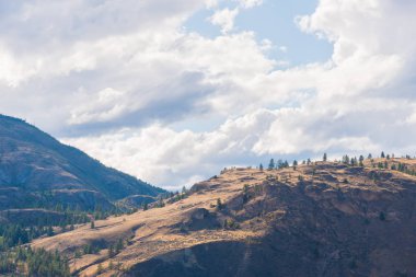Okanagan Vadisi'nde karamsar bulutlar ve mavi gökyüzü ile güneş ışığı altında parlayan uzak tepeler
