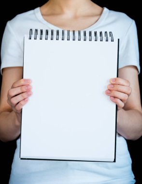 Siyah arka planda beyaz tişört giyen bir kadının tuttuğu boş not defterinin yakın plan görüntüsü.