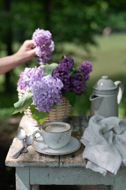Bahçede kahvaltı: eclairs, kahve fincanı, kahve demliği, bir sepet içinde leylak çiçekler.
