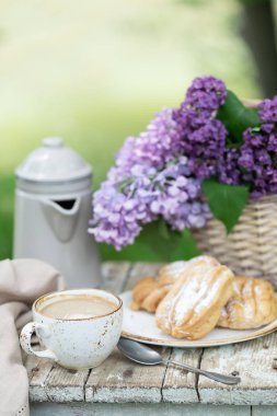 Bahçede kahvaltı: eclairs, kahve fincanı, kahve demliği, bir sepet içinde leylak çiçekler.