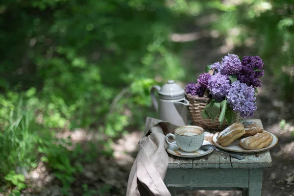 Petit déjeuner dans le jardin : éclairs, tasse de café, cafetière, fleurs de lilas dans un panier . — Photo