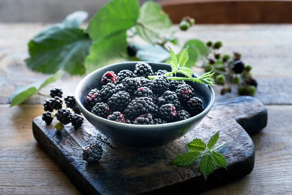 Olgun böğürtlen seramik bir kapta hasat edilir. Yeşil yaprakları ve dağınık çilek. Ahşap arka plan