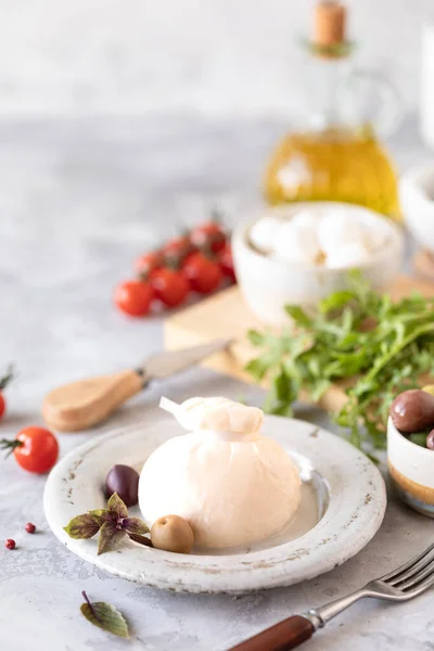 Quesos italianos burrata y mozzarella con aceitunas, tomates cherry y rúcula sobre un plato redondo blanco sobre un fondo claro —  Fotos de Stock