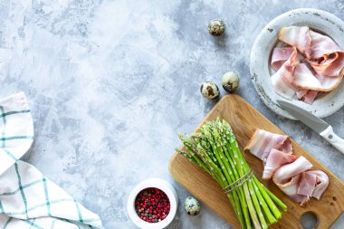 Gri beton arka planda yemek pişirmek için gerekli malzemeler. Bir demet taze kuşkonmaz, yumurta ve pastırma. Üst Manzara. Uzayı kopyala Yatay