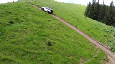 Dağlık bir arazide toprak bir yolda beyaz bir SUV sürüyor. Hava görünümü.