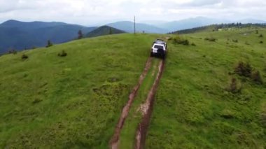 Dağlık bir arazide toprak bir yolda beyaz bir SUV sürüyor. Hava görünümü.