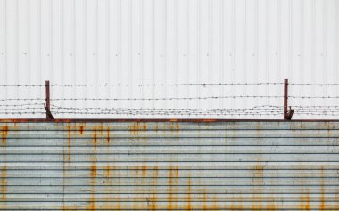 Paslı parantezler üzerine gerilmiş dikenli tel, bulutlu bir gökyüzüne karşı metal levhalardan yapılmış bir çite monte edilmiş..