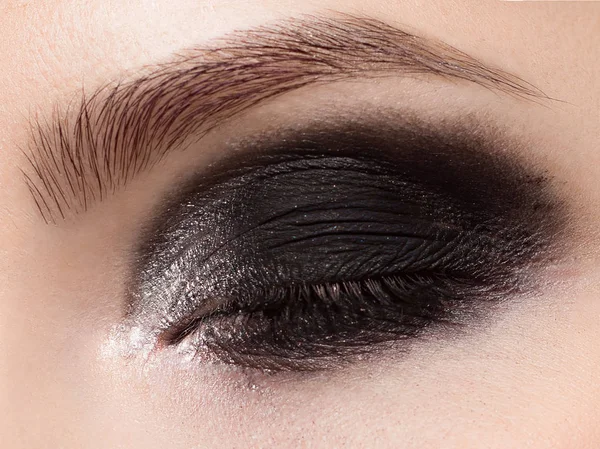 Macro photo of a closed eye with smokey black shadows, long eyelashes and thick even eyebrows. Perfect fashionable evening eye makeup. The beauty of classic make-up. White glitter