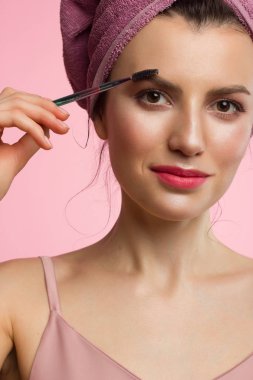 Doğal makyajlı ve temiz tenli, kafasında havlu olan güzel bir kadının yakın çekim. Kız kalın kaşları tarıyor ve kameraya bakıyor. Kozmetoloji ve spa