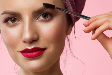 Doğal makyajlı ve temiz tenli, kafasında havlu olan güzel bir kadının yakın çekim. Pembe tombul dudaklar. Kız kalın kaşları tarıyor ve kameraya bakıyor. Kozmetoloji ve spa