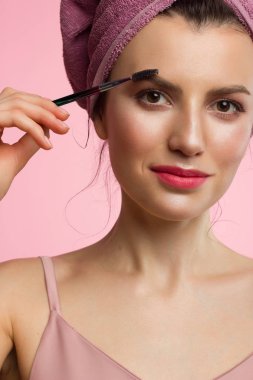 Doğal makyajlı ve temiz tenli, kafasında havlu olan güzel bir kadının yakın çekim. Kız kalın kaşları tarıyor ve kameraya bakıyor. Kozmetoloji ve spa
