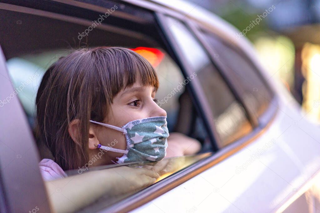a Caucasian little girl in a  mask looking out of the open window of a car. Hygiene mask protection from a virus, air pollution concepts