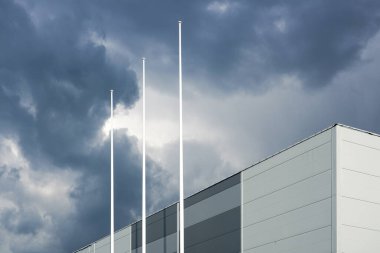 Yeni yapım sandviç paneli cephesi ve arka planda gri fırtına bulutları üzerinde bayrakları olmayan üç bayrak direği.