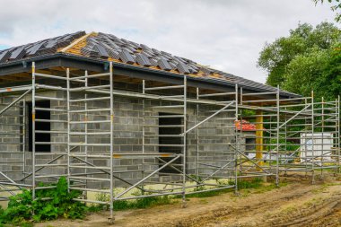 İskeleli ve kısmen döşenmiş çatı kiremitleri olan tamamlanmamış tuğla ev. Yerleşim alanı çatı ve duvar inşaatının ilerlediğini gösteriyor.