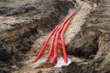 Parlak kırmızı borular ve kalın elektrik kabloları bir inşaat alanındaki kumlu bir siperden geçiyor ve suyla dolu bir alanda bitiyor.