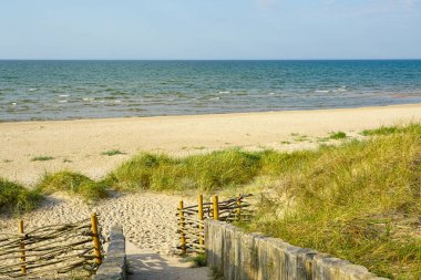 Kıyı kum tepeleri boyunca uzanan ahşap bir yolun manzarası, açık yaz gökyüzü altında kumlu bir Baltık sahili ve yumuşak deniz dalgalarına açılıyor.