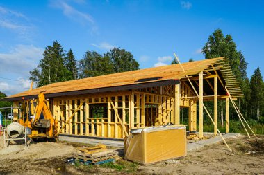 ICF temelinde çatı kurulumu olan ahşap ev iskeleti. Güneşli inşaat alanında kazıcı, araç gereç ve inşaat malzemeleri