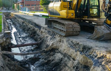 Sarı bir kazıcı, görünür borular, toprak katmanları ve altyapı onarımı için çamurlu su içeren bir hendek kazan kentsel bir şantiyede çalışıyor.