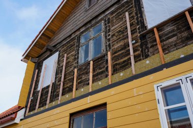 Yeni sarı kaplama, yalıtım ve pencere güncellemeleri ile yenilenen eski ahşap bir evin yakın çekimi.