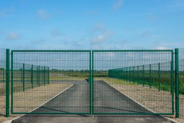 Kırsal bölgedeki özel asfalt yolu çevreleyen çitli yeşil metal güvenlik kapısı erişim kontrolünü, sınırı ve kısıtlı girişi sembolize ediyor.