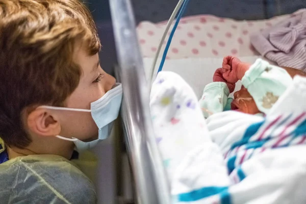 Young boy big brother wearing face mask and gown while looking at his newborn sister at a hospital nursery