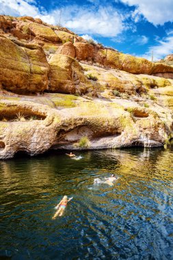Unidentifiable people swimming in Canyon Lake, Arizona to reach the rocky mountain shore to go cliff diving clipart