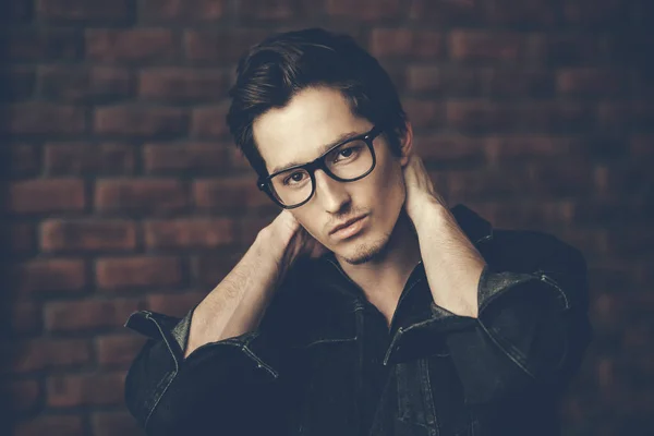 Male beauty, fashion. Handsome young man posing in spectacles and black  denim jacket by the brick wall. Hair styling. - Stock Image - Everypixel
