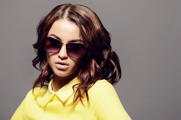 Portrait of a pretty woman in yellow blouse and sunglasses over gray background. Fashion shot.