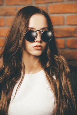 Şapka ve güneş gözlüğü çekici genç bir kadın portresi yakın çekim bir tuğla duvar tarafından duruyor. Stüdyo vurdu. Güzellik, moda kavramı.