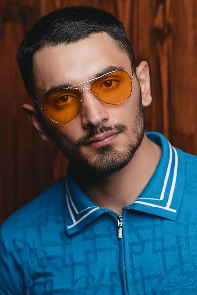 Vogue shot of a handsome male model in a T-shirt and yellow sunglasses standing by a wooden wall. Men\'s beauty, fashion.