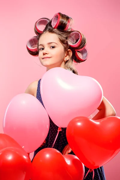 Portrait of a pin-up little girl with curlers in her hair and balloons. Studio shot over pink background. Kid\'s fashion.