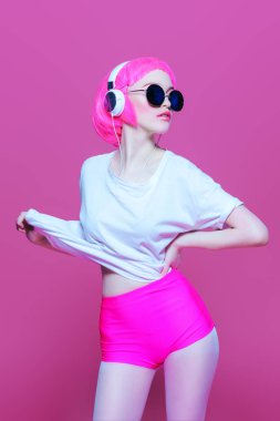 Trendy kız pembe saçlı güneş gözlüğü takıyor kulaklıklar müzik hoşlanır. Pembe bir arka plan. Gençlik stil, eğlence.