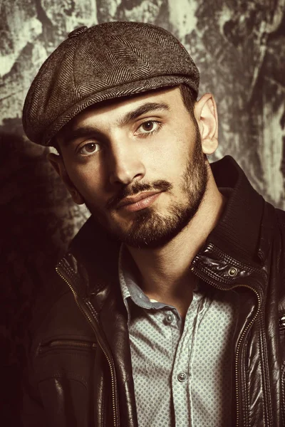 Portrait of a handsome man in a cap and leather jacket on a grunge background. Studio shot. Men's beauty, fashion. Men's barbershop.