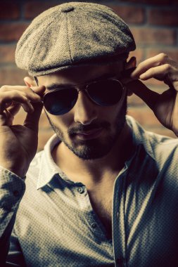 Bir kap ve tuğla duvar arka plan üzerinde güneş gözlüğü yakışıklı bir adam portresi. Erkek güzelliği, moda. Erkek berber.
