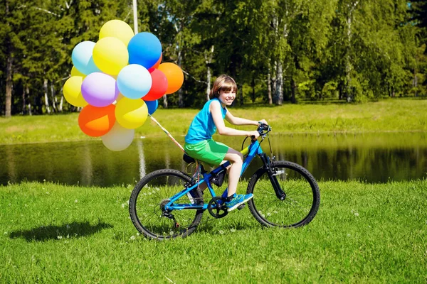 Happy boy is riding a bicycle with colored balloons in a park. Summer holidays. Birthday.