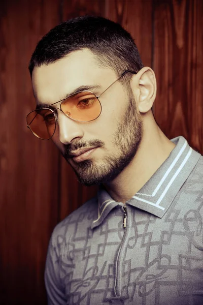 Vogue shot of a handsome male model in a T-shirt and yellow sunglasses standing by a wooden wall. Men\'s beauty, fashion.