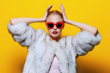 Çok güzel sarışın kadın lüks kürk ceket ve pin-up güneş gözlüğü içinde poz. Sarı arka plan. Moda, güzellik. Stüdyo vurdu.