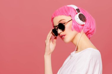 Trendy kız pembe saçlı güneş gözlüğü takıyor kulaklıklar müzik hoşlanır. Pembe bir arka plan. Gençlik stil, eğlence.