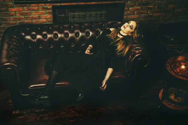 Magnificent woman in black gothic dress lies on a leather sofa. Fashion. Gothic style.