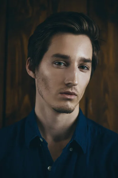 Vogue shot of a handsome male model in a denim shirt standing by a wooden wall. Men\'s beauty, fashion.