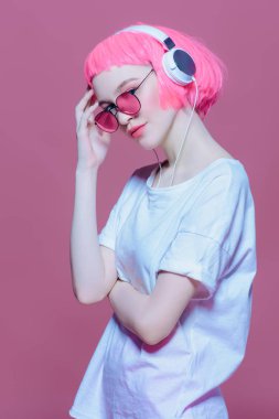 Trendy kız pembe saçlı güneş gözlüğü takıyor kulaklıklar müzik hoşlanır. Pembe bir arka plan. Gençlik stil, eğlence.