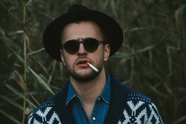 Yakın çekim portre hırka ve şapka açık sonbahar doğa arka plan üzerinde sigara yakışıklı acımasız bir adam. Sonbahar moda. Erkek moda. 